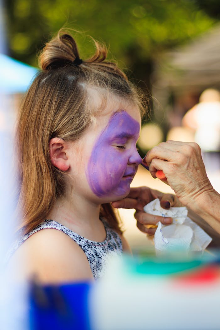 services-03 A young girl with blond hair is getting her face painted purple during an outdoor event on a sunny day.